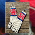 A pair of woven fringe earrings with pink white, and blue woven patterns, displayed on a wooden surface with a blurred background of colorful yarn balls.