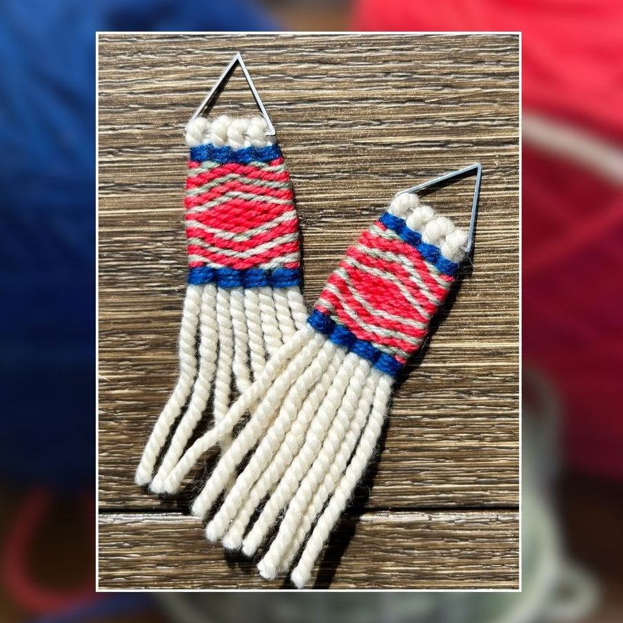 A pair of woven fringe earrings with pink white, and blue woven patterns, displayed on a wooden surface with a blurred background of colorful yarn balls.