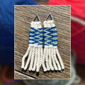 A pair of handmade woven fringe earrings with geometric blue, white, and light green patterns, displayed on a wooden surface with a blurred background of colorful yarn balls.