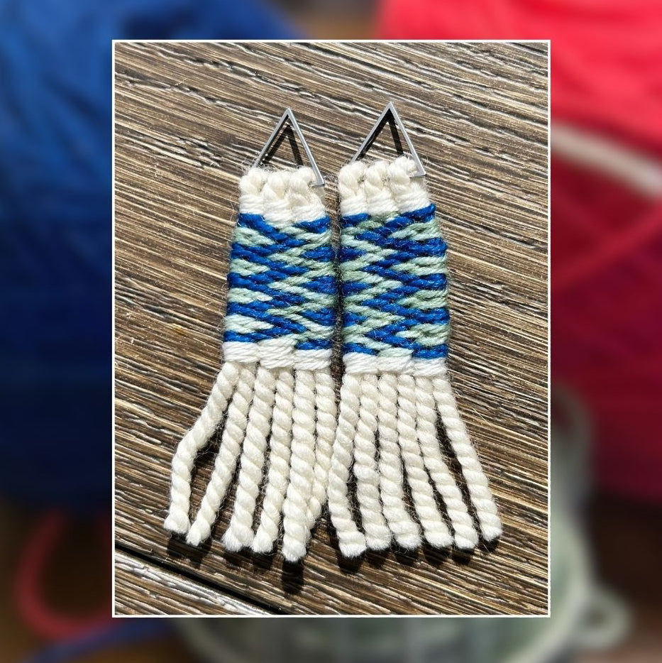 A pair of handmade woven fringe earrings with geometric blue, white, and light green patterns, displayed on a wooden surface with a blurred background of colorful yarn balls.