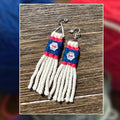A pair of handmade earrings with cream-colored warp, blue and pink wrapped sections, and a small white floral detail, displayed on a wooden surface with a blurred background of colorful yarn balls.