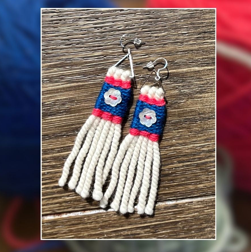 A pair of handmade earrings with cream-colored warp, blue and pink wrapped sections, and a small white floral detail, displayed on a wooden surface with a blurred background of colorful yarn balls.
