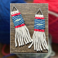 A pair of handmade woven earrings with geometric blue, white, and pink patterns, displayed on a wooden surface with a blurred background of colorful yarn balls.
