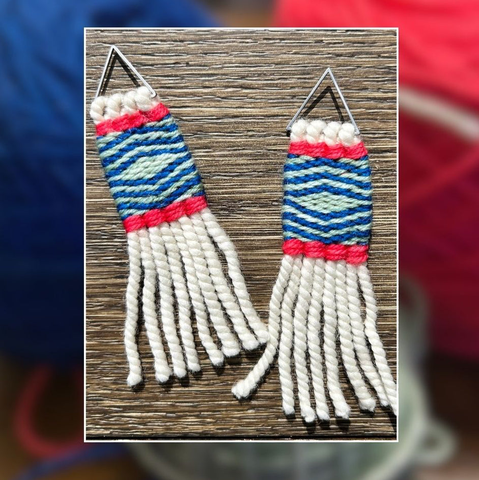 A pair of handmade woven earrings with geometric blue, white, and pink patterns, displayed on a wooden surface with a blurred background of colorful yarn balls.