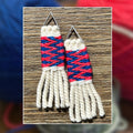 A pair of handmade woven earrings with triangular silver metal tops, featuring cream fringe, a pink and blue woven band, and a white woven top section, displayed on a wooden surface with a blurred background of colorful yarn balls.