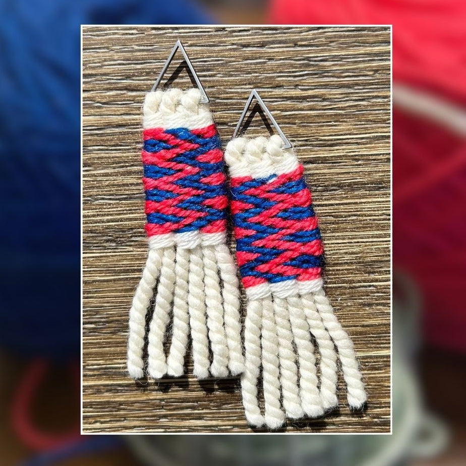 A pair of handmade woven earrings with triangular silver metal tops, featuring cream fringe, a pink and blue woven band, and a white woven top section, displayed on a wooden surface with a blurred background of colorful yarn balls.