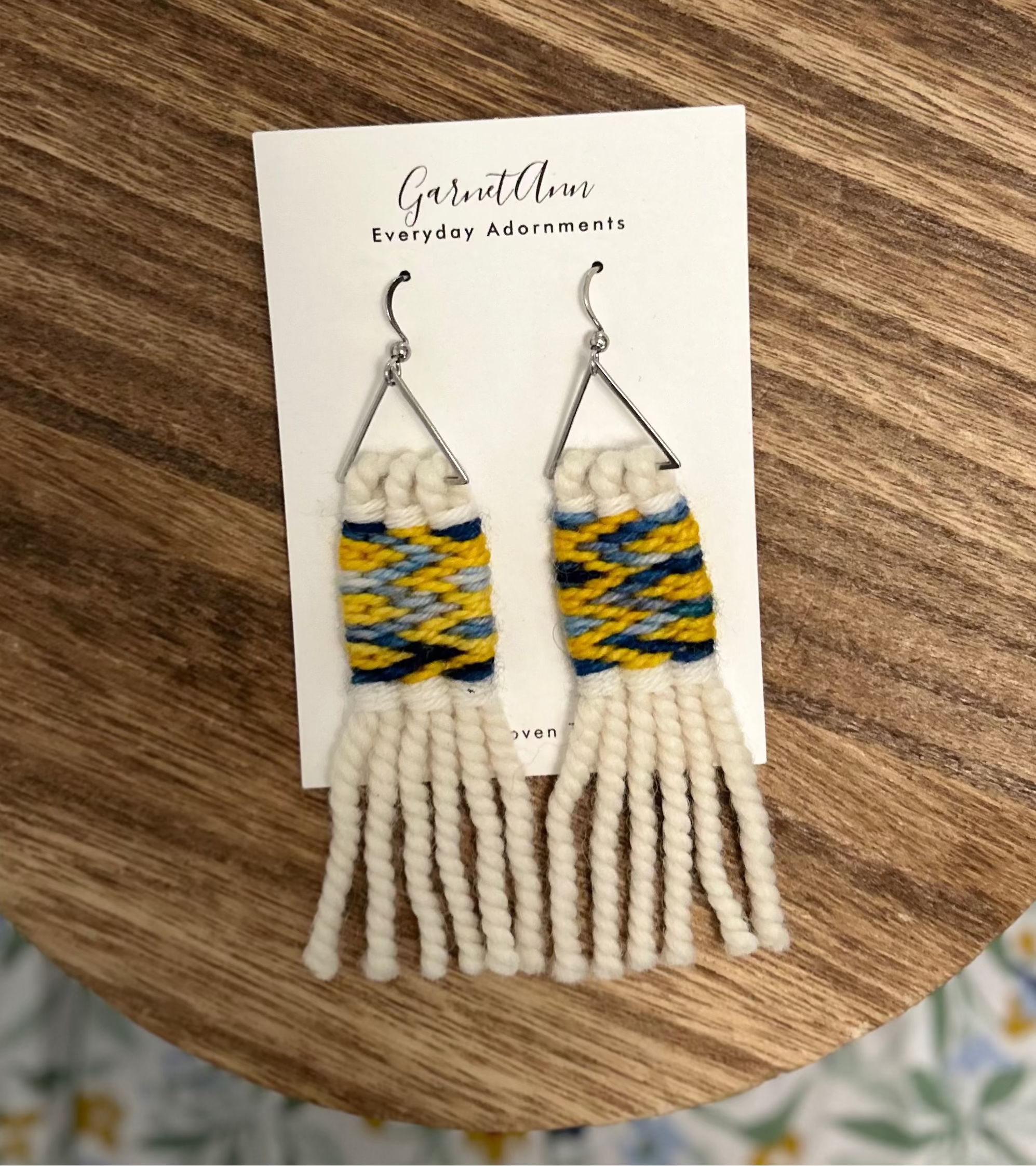 A pair of handmade woven fringe earrings displayed on a white card, placed on a round wooden coaster over a floral fabric background. The earrings feature silver-tone hooks, yellow and blue woven yarn, and white warps.
